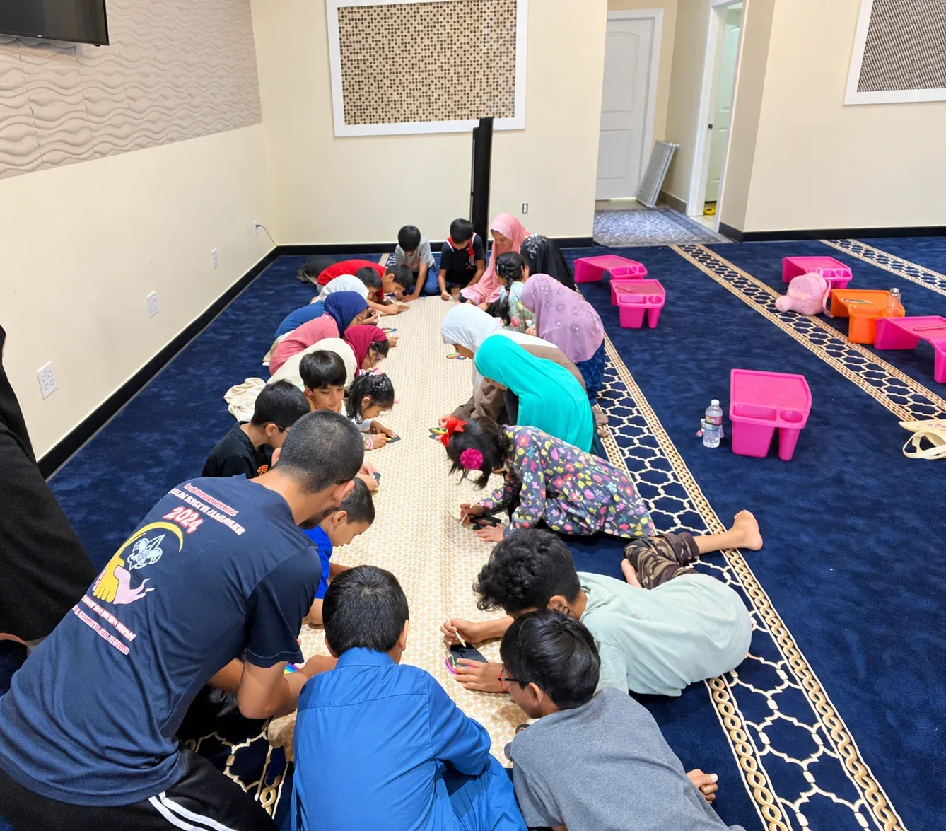 Children bent over crafts along a strip of paper on the masjid carpet